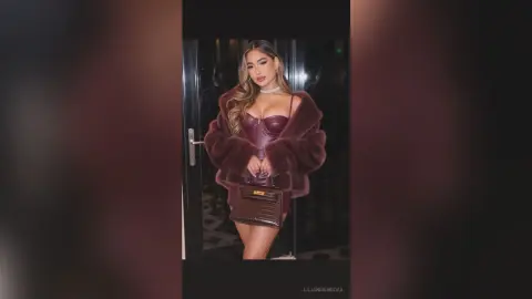 Glamorous woman in a burgundy faux fur coat and corset outfit posing elegantly in a dimly lit doorway.