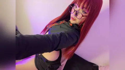 Red-haired woman in a black long-sleeve shirt and glasses posing in soft pink lighting.