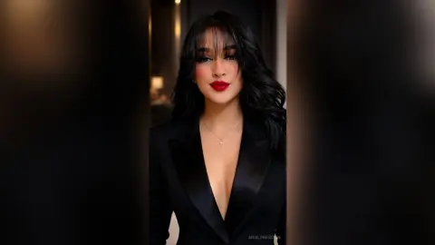 Elegant woman with long dark hair and red lipstick wearing a chic black satin blazer in a moody setting.