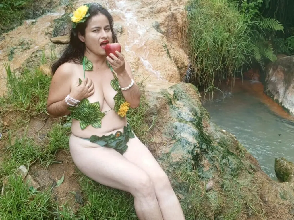 Woman in a leafy green nature goddess outfit sitting by a jungle waterfall in a mystical forest setting.