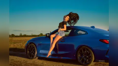 Fashionable woman in a black top and denim shorts leaning on a blue car in a sunny field at sunset.