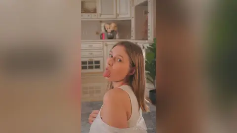 Playful young woman in a white tank top making a funny face in a bright, modern kitchen setting.