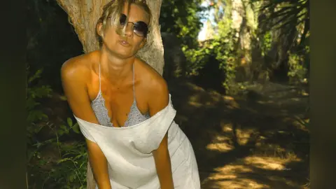 AtlamaDesmond poses outdoors against a tree, wearing a white tank top and sunglasses with her blonde hair down.
