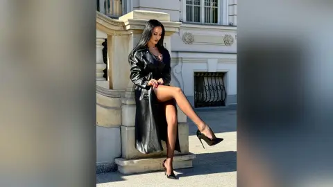Stylish woman in a long black leather coat and high heels posing outdoors by a classic stone building.