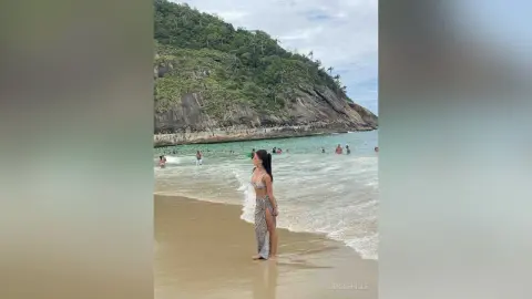 Young woman in a patterned bikini and long skirt walking on a tropical beach with ocean waves and cliffs.