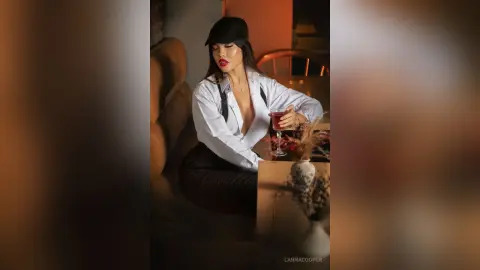 Stylish woman in a white shirt and black cap sipping red wine in a moody, dimly lit lounge setting.