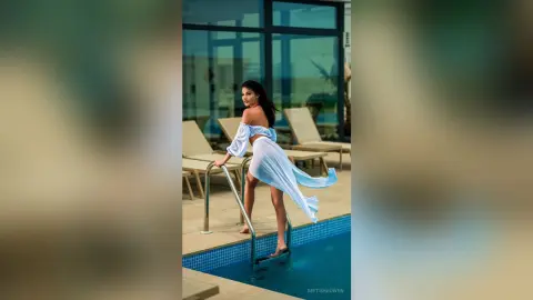 Elegant woman in a flowing white off-the-shoulder dress posing by a luxury resort swimming pool.