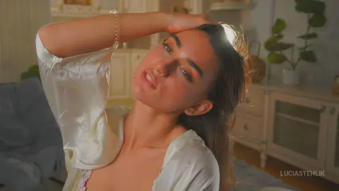 Young woman in a white silk pajama top relaxing in a sunlit bedroom with a dreamy, peaceful mood.