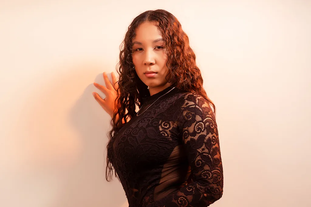 EyshelNoah poses with her hands up against a plain white wall, wearing a black lace long-sleeve top and letting her long wavy hair hang down.