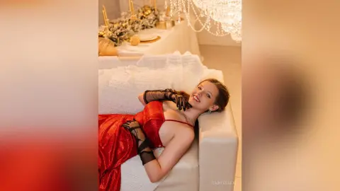 Smiling woman in a glamorous red sequin dress and black lace gloves relaxing on a white sofa in a festive setting.