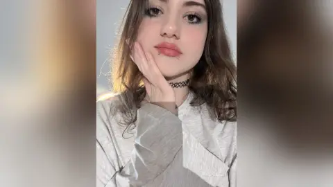 AriaLush poses with her hand against her cheek, wearing a grey long-sleeve shirt and a black choker, showing her long wavy brown hair and lip piercing.