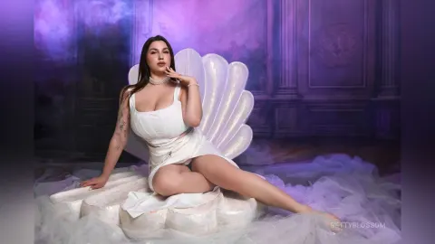 BettyBlossom — BettyBlossom sits on a large white seashell chair wearing a white crop top and skirt against a purple background.