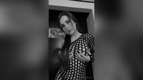 Moody black and white portrait of a woman in a black polka dot sheer dress indoors.