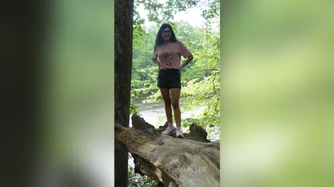 Young woman in a pink t-shirt and black shorts standing on a fallen log in a lush green forest setting.
