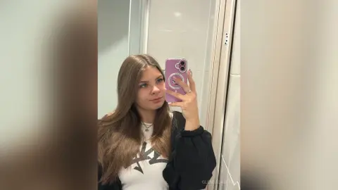 Young woman with long brown hair taking a mirror selfie in a white tiled bathroom wearing a casual graphic tee.