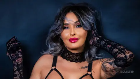 Glamorous woman with wavy grey hair wearing black lace gloves and lingerie against a dark moody background.
