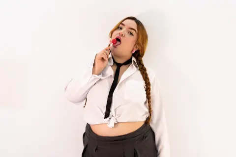 Young woman in a white shirt and black tie eating a red lollipop against a white background.