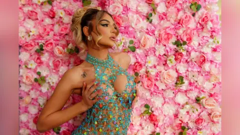 Anastasia poses against a wall of pink roses wearing a sheer, sparkling turquoise mesh bodysuit.