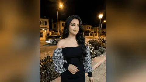 Young woman in a black off-the-shoulder dress posing outdoors at night under warm streetlights.