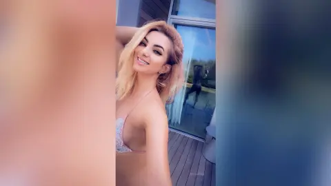 FelishaRose smiles while posing in a light pink lace bra against a glass window or door.