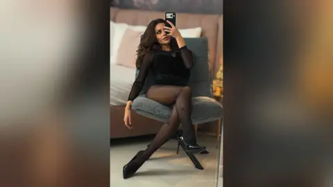 AnaMathers poses sitting on a grey chair wearing a black long-sleeve bodysuit and sheer black stockings with small star details.