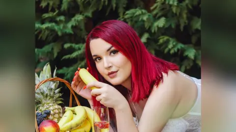 Red-haired woman in a white lace top holding a banana near a fruit basket in a lush green garden setting.