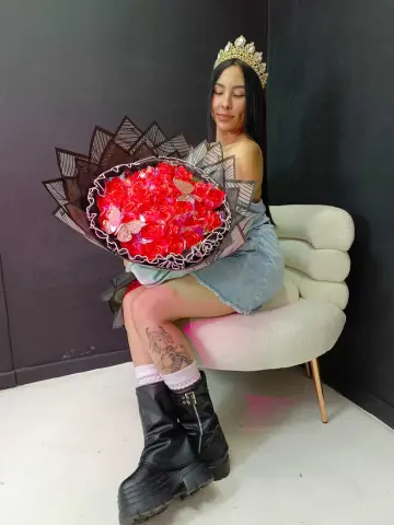 MorganHarper wears a gold crown and a denim skirt while sitting on a white chair, holding a large bouquet of red roses.