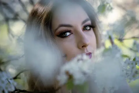 Blonde woman with winged eyeliner peeking through white spring blossoms in a dreamy, soft-focus garden setting.