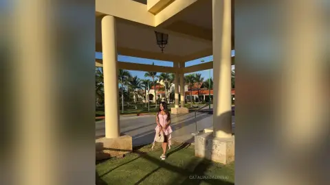 Elegant woman in a pink dress standing under a sunlit terrace at a tropical resort during golden hour.