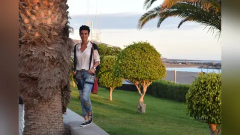 Stylish woman in white blouse and denim jeans standing in a sunny tropical garden near a beach.