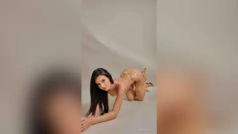 JasmineHille poses on all fours against a plain background, wearing a gold string bikini that shows off her long dark hair and bare skin.