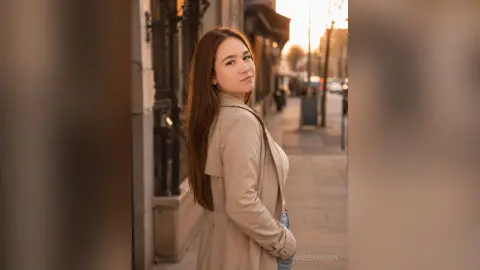 Smiling young woman in a beige trench coat walking on a sunny city street during golden hour.