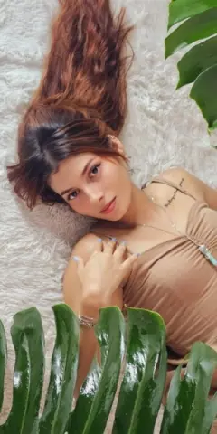 Young woman with long brown hair in a tan tank top lying on a white shag rug near green tropical leaves.
