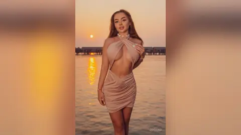 Beautiful woman in a beige halter dress posing by the ocean during a golden sunset.