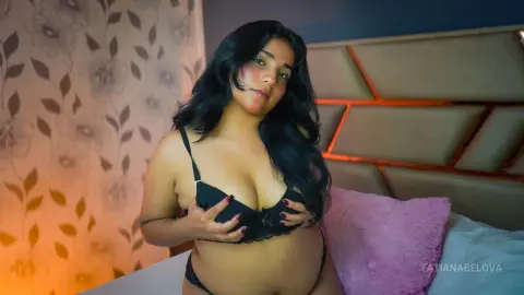 Young woman in black lace lingerie relaxing on a bed with floral wallpaper in a cozy bedroom setting.