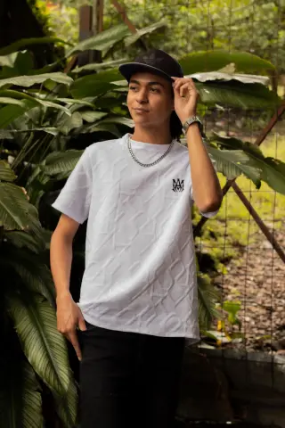 KylePresly wears a white t-shirt and black cap while standing outdoors near large green leaves.