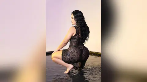 MaraGrief poses in a squatting position wearing a sheer black lace bodysuit while standing in shallow water against a plain light background.