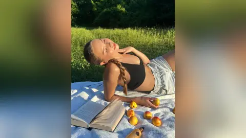KrystalLaaz lies on a white blanket in a grassy field, wearing a black crop top and denim shorts while relaxing in the sun.