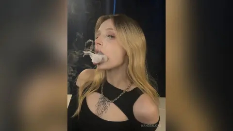 Blonde woman in a black cutout top exhaling smoke in a moody, dark indoor setting.
