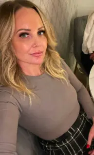Blonde woman in a grey long-sleeve top and plaid skirt sitting in a cozy indoor setting.