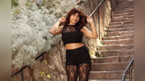 Young woman in a black lace crop top and shorts posing on stone stairs amidst white flowering bushes.