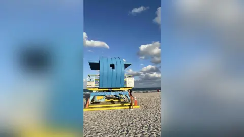 A woman in a black bikini relaxing on a sunny Miami Beach sand near a blue lifeguard tower under a clear sky.
