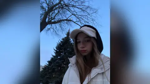 MagalyBoffa wearing a white hooded jacket and a white furry hat, posing outdoors against a blue sky and bare tree branches.