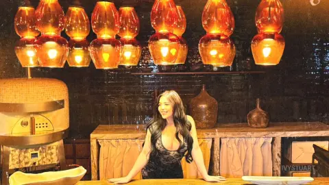 Smiling woman in a black dress sitting at a rustic wooden bar with warm glowing amber pendant lights.