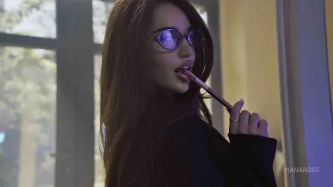 Young woman with glasses and dark hair wearing a black outfit, posing thoughtfully in soft indoor lighting.