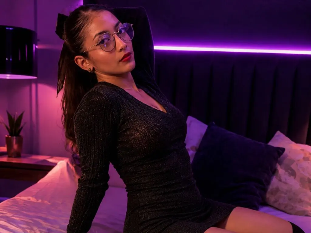 Young woman in a sparkly black long-sleeve dress sitting on a bed in a moody, purple-lit bedroom.