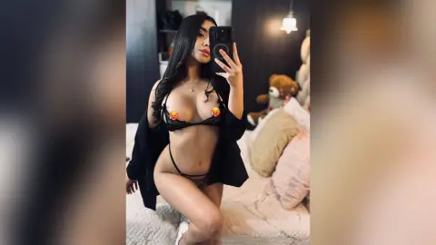 Young woman in black lingerie and an open cardigan taking a mirror selfie in a cozy bedroom setting.