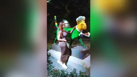 NohomiCambel stands next to a large green cat statue wearing a brown sleeveless dress and white sneakers.