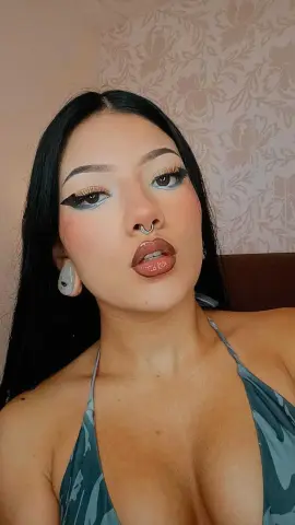 Young woman with long dark hair and winged eyeliner wearing a grey camo halter top in a cozy room.