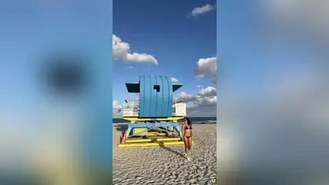 Woman in black bikini walking on Miami Beach sand near a blue lifeguard tower under a sunny sky.